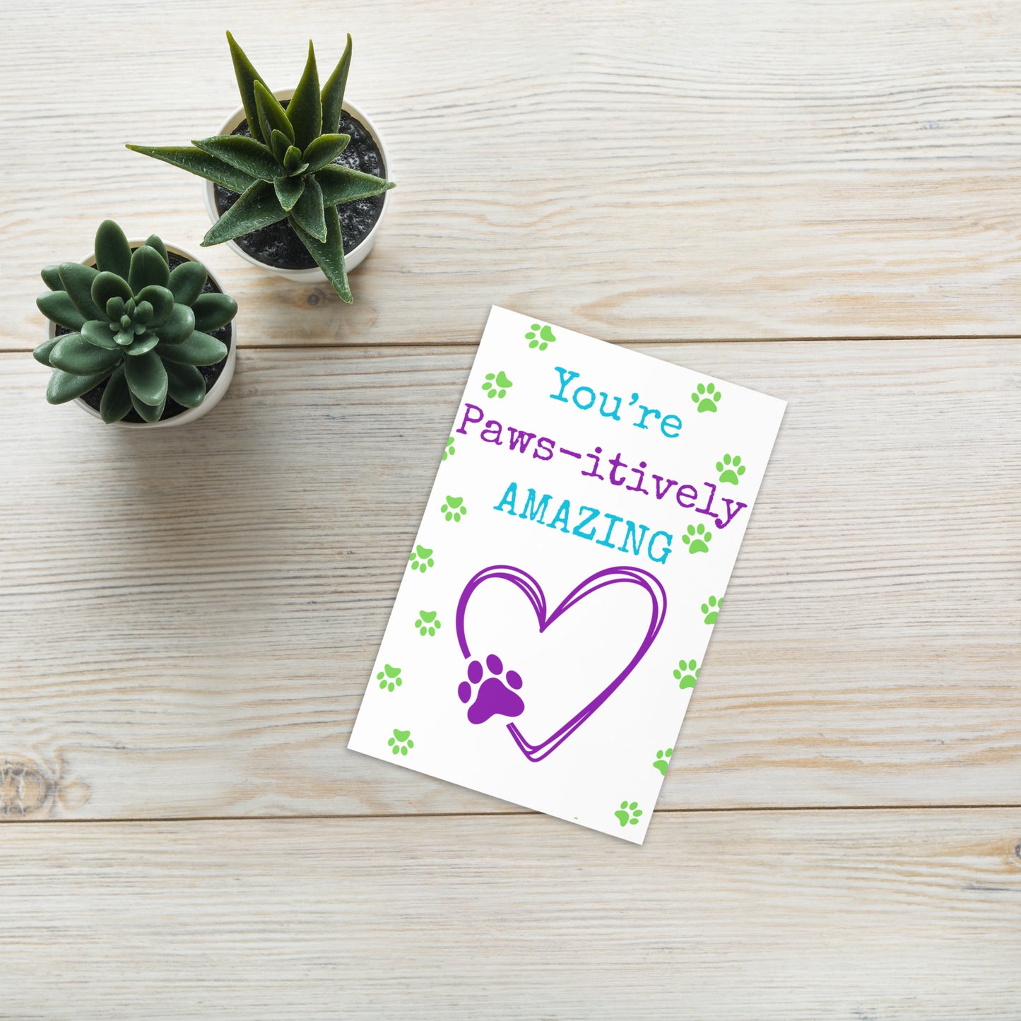 Card with 'You're Paws-itively Amazing' text on a wooden surface with plants.